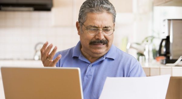 Image is of an older latino man sitting at his laptop with a piece of paper looking annoyed at it
